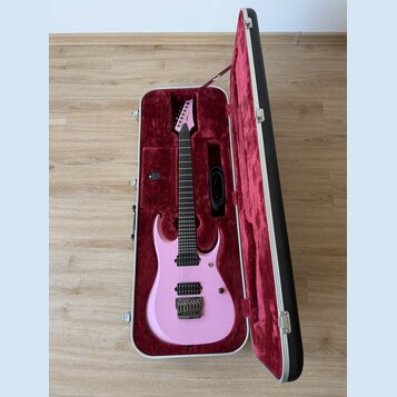 Růžová elektrická kytara v tvrdém kufru s červeným polstrováním. Na hlavě hmatníku je bílý nápis Ibanez. Kytara má dva humbuckery, Tremolo most a čer…
