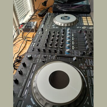 Černý DJ 컨트롤러 s ovládacími prvky, posuvníky a dvěma stříbrnými jog-wheels. Na povrchu je viditelný nápis Pioneer DJ. Zařízení vykazuje běžné známky p…