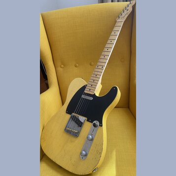 Žlutá elektrická kytara s tvarem typu Telecaster a černým pickguardem. Na hlavě hředu je viditelné logo Fender. Tělo kytary vykazuje výrazné opotřebe…