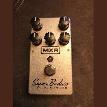 Stříbrný efekt pedál s pěti černými ovládacími knoflíky. Na přední straně je umístěno logo MXR, nápis Super Badass a pod ním slovo DISTORTION. V horn…