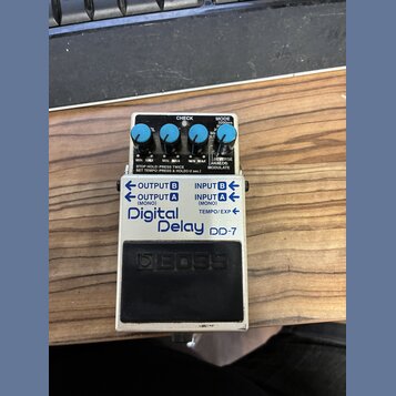 Stompbox efekt s bílým kovovým šasi a černým spínacím pedálem. Na přední straně je nápis Digital Delay DD-7 a logo BOSS. Zařízení má čtyři modré ovlá…
