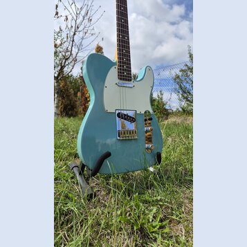 Světle modrá elektrická kytara typu Telecaster s bílým pickguardem a hnedým hmatníkem. Nástroj je umístěn na černém stojanu v trávě. Na kytare nejsou…