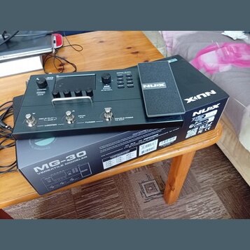 Černý kytarový procesor NUX MG-30 položený na své originální krabici s nápisem MG-30 VERSATILE MODELER. Zařízení má digitální displej, několik ovláda…