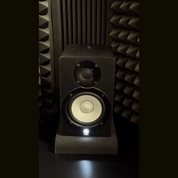 Černý studiový monitor v hranaté skříni, umístěný na černém podstavci. Reproduktor má v horní části malý výšek s mřížkou a ve spodní části větší bílý…