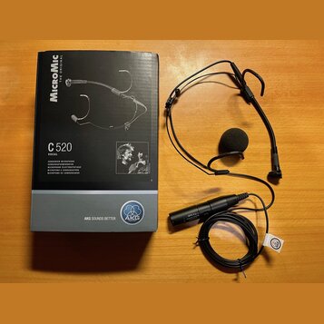 Na fotografii je černý hlavový mikrofon s černou molovou větřnicí a doprovodná krabice. Na krabici jsou nápisy MICROMIC THE ORIGINAL, C 520 VOCAL a l…