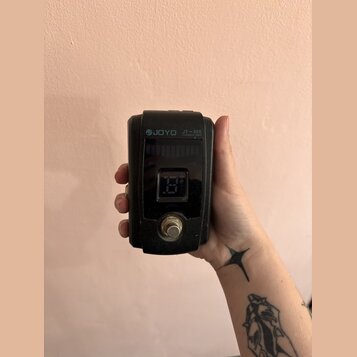 Černý ladicí pedál s digitálním displejem. Na horní části je bílým nápisem značka JOYO a model JT-305 CHROMATIC TUNER. Povrch zařízení vykazuje drobn…
