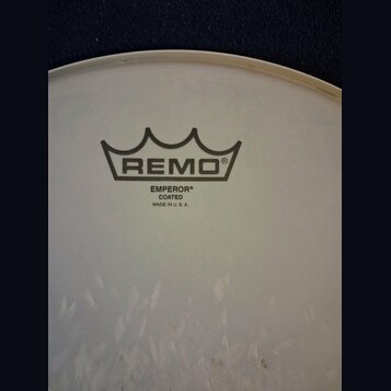 Detail bílého, matného bicího koženu. Uprostřed je černé logo REMO v podobě koruny a pod ním nápisy EMPEROR®, COATED a MADE IN U. S. A. V dolní části…