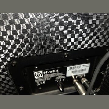 Detail zadní strany reproduktoru s černým šachovnicovým potahem. Na černé plošce je bílý nápis PF-115HE a logo s názvem Ampeg. Jsou zde uvedeny speci…