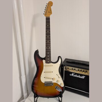 Elektrická kytara s tónem sunburst, bílým pickguardem a javorovou hlavou. Na hlavě je viditelné logo Fender. Vedle kytary stojí černý kytarový zesilo…