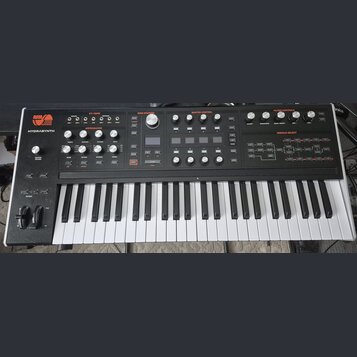 Černý synthesizér s bílými a černými klávesami. V levém horním rohu je logo ASM ASHLEY VOLUME MACHINES a nápis HYDRASYNTH. Panel obsahuje množství ov…