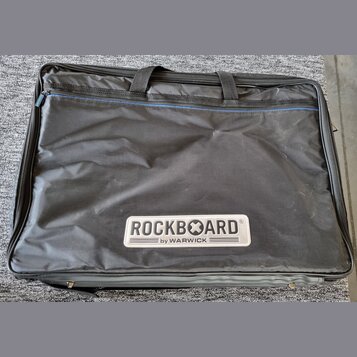 Černý textilní pouzdro na hudební nástroj s dvěma rukojtěmi a zipem. V dolní části je bílý obdélníkový štítek s nápisem ROCKBOARD® a pod ním menší te…