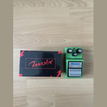 Zelený efekt pedál s kovovým šasi a stříbrným spínacím tlačítkem. Na pedálu je bílý štítek s nápisy Maxon, OD-9 a Overdrive. Vedle pedálu je černá kr…