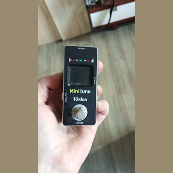 Černý kytarový ladič v kompaktním kovovém provedení. Na přední straně je displej, tři kontrolky a nápisy MiniTune a Vivlex. V horní části je označení…