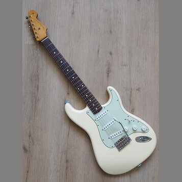 Bílá elektrická kytara s jasně viditelným logem Fender na hlavě. Nástroj má tmavý hmatník, světle zelený pickguard a tři snímače. Povrch kytary vypad…