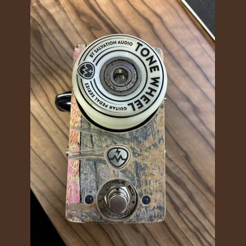 Kytarový efekt s bílým ovládacím knoflíkem a šasi z texturovaného materiálu v hnědo-šedých tónech. Na knoflíku je nápis BY SALVATION AUDIO TONE WHEEL…