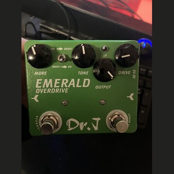 Zelený kytarový efekt s nápisy EMERALD OVERDRIVE a Dr. J. Na horní straně jsou čtyři černé otočné knoflíky s popisky MORE, TONE, OUTPUT a DRIVE. Mezi…