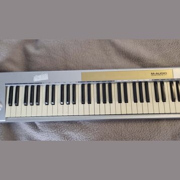 Stříbrný MIDI kontrolér s klávesnicí. Na horním panelu je zlatý pruh s nápisem M-AUDIO KEYSTATION 61mkII. Bílé klávesy vykazují zežlutnutí, na stříbr…