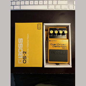 Žlutý kytarový efekt BOSS OS-2 Over Drive/Distortion v originální žluté krabici. Na pedálu jsou čtyři ovládací knofíky a nápisy CHECK, TONE, DRIVE a…