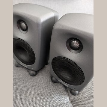 Dva šedé studiové monitory s matným povrchem a zaoblenými hranami. Na spodní části obou reproduktorů je bílý nápis GENELEC. Každý monitor má v horní…