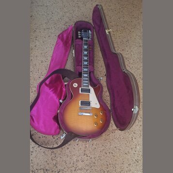 Elektrická kytara s tónem sunburst v hnědých a žlutých odstínech, bílým pickguardem a dvěma humbuckery. Nástroj je umístěn v otevřeném hnědém tvrdém…