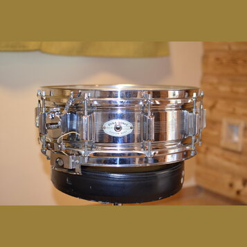 Kovový snare drum v chromovaném provedení. Na korpusu je vidět oválný štítek s nápisy DYNA-SONIC, SERIAL No. (číslo nečitelné) a CUSTOM BUILT. Na mec…