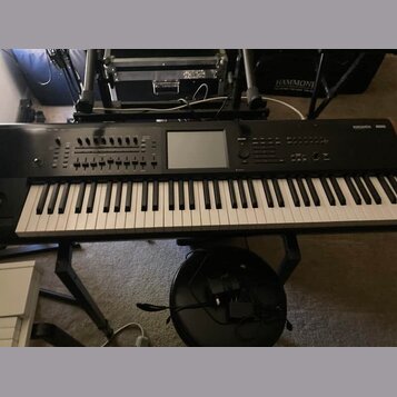Černý digitální klavír umístěný na černém stojanu. V pravém horním rohu je bílý nápis KRONOS Xsynth. Nástroj má v horní části ovládací prvky, tlačítk…