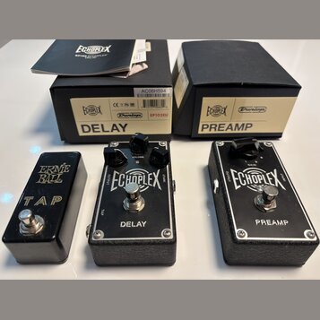 Tři černé kytarové efekty v kovovém provedení s texturovaným spodkem. První pedál má nápisy ERNIE BALL a TAP, prostřední nápisy ECHOPLEX a DELAY s ov…