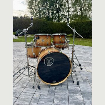 Akustická bicí souprava z přírodního dřeva s kovovým hardwarem. Na basovém bubnu je černé logo s nápisem „SINCE 2003 DC DRUMS handcrafted in germany“…