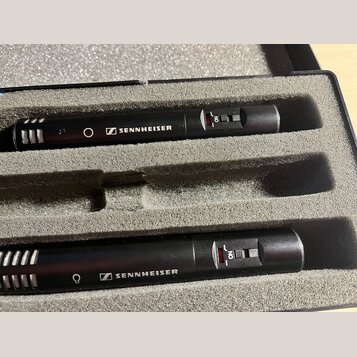 Dvě černá kondenzátorová mikrofony umístěná v šedém pěnovém pouzdře. Na obou je bílý nápis SENNHEISER s doprovodným logem a červený přepínač s nápise…