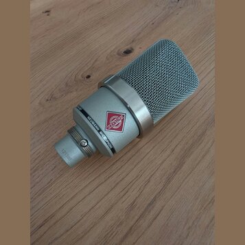 Stříbrný kondenzátorový mikrofon s kovovou síťovkou. Na těle je nápis NEUMANN MICROPHONE a červenobílý štítek s nečitelným nápisem. Na spodní části j…