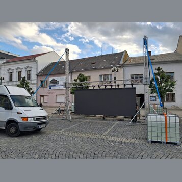 Fotografie zachycuje venkovní instalaci kovového hliníkového truss systému s černou plátnem v pozadí. Konstrukce je zajištěna modrými napínacími popr…