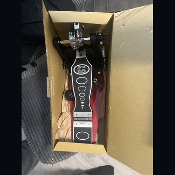 V kartónové krabici se nachází červený bubnový pedál. Na horní části je bílý oválný štítek s nápisem DRUMS STAND a pod ním Stabla. Pedál má kovové me…