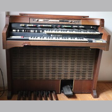 Hnědý dřevěný orgán se dvěma klávesnicemi a pedálovým hracím polem. Na spodní klaviatuře je umístěn nápis HAMMOND. Horní panel obsahuje řadu bílých a…