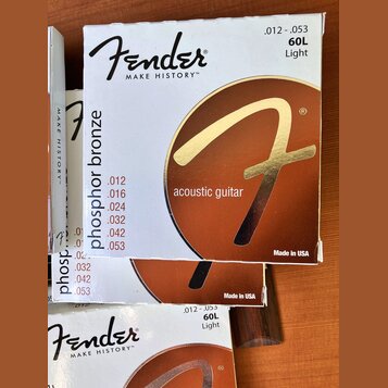 Fotografie zobrazuje tři krabičky strun pro akustickou kytaru. Na obalech je bílé pozadí, hnědý kruh se zlatým logem „F“ a nápisy „Fender“, „MAKE HIS…