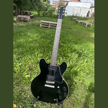 Černá elektrická kytara s polodupatým tělem a bílým lemováním. Na hlavě hředu je viditelné logo Gibson. Nástroj má dva humbuckery, čtyři ovládací kno…