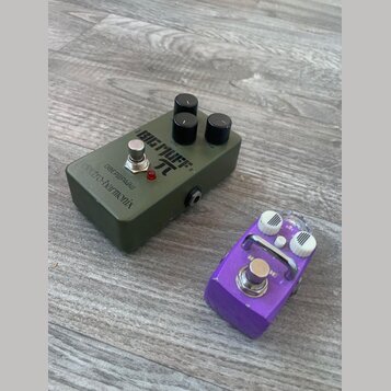 Na fotografii jsou dva efektové pedály na dřevěné podlaze. Větší pedál je olivově zelený s nápisy BIG MUFF π, electro-harmonix a ОВЕРДРАЙВ. Menší ped…