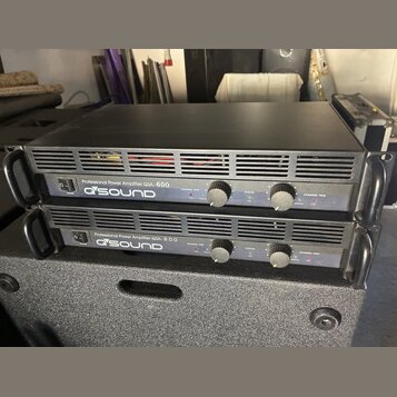 Dvě černé rackové konce s nápisem Professional Power Amplifier QSA-600 a logem QSOUND. Každý přístroj má v přední části vypínač, dva ovratové ovládac…