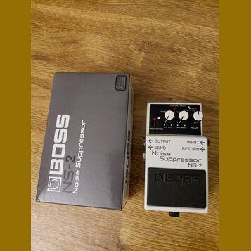 Na fotografii je kytarový pedál Boss Noise Suppressor NS-2 v bílém provedení s černým spínacím tlačítkem. Vedle pedálu je umístěna jeho originální še…