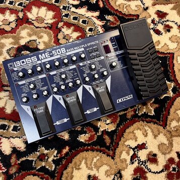Modrý multifunkční efektový procesor s nápisem BOSS ME-50B BASS MULTIPLE EFFECTS a logem COSM. Zařízení obsahuje několik ovládacích knoflíků, přepína…