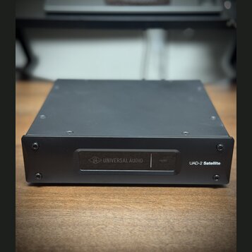 Černý kovový přístroj obdélníkového tvaru. Na přední straně je nápis UAD-2 Satellite a panel s logem a názvem UNIVERSAL AUDIO oraz nápisem HOST. Povr…
