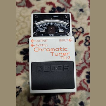 Bílý kytarový ladič značky BOSS, model Chromatic Tuner TU-3. Na horní straně je analogový displej s nápisy CHECK, PITCH a označením tónů. V horní čás…