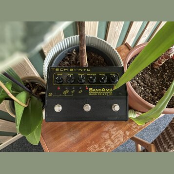 Černý kytarový pedál s nápisem TECH 21 NYC a SansAmp PROGRAMMABLE BASS DRIVER DI. Horní panel obsahuje šest otočných potenciometrů s popisky DRIVE, B…