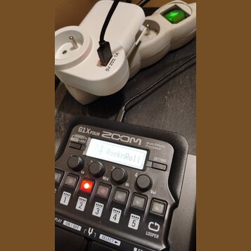 Černý kytarový procesor s nápisy ZOOM G1X FOUR a Multi-Effects Processor. Na displeji je zobrazen text 24 RocknRoll. Zařízení je připojeno k bílému n…