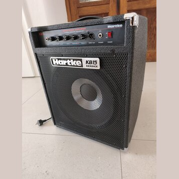 Černý basový kombozesilovač s kovovou ochrannou mřížkou a hliníkovými rohy. Na předním panelu je bílý nápis Hartke KB15 KICKBACK a v horní části nápi…