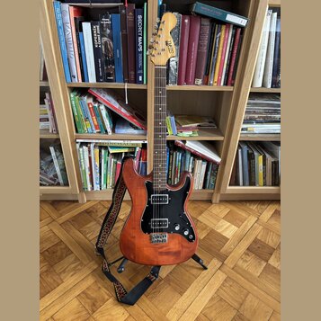 Elektrická kytara s dřevem v přirozené hnědé barvě, černým pickguardem a černými potenciometry. Na hlavě nástroje je svisle napsané logo Squier. Kyta…