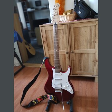 červená elektrická kytara s bílým pickguardem a hnedým hmatníkem. Na hlavě je viditelný nápis YAMAHA. Kytara je umístěna na stojanu a má připojený če…
