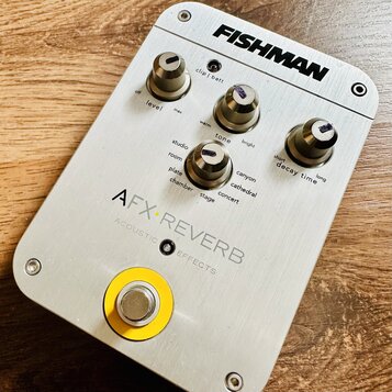 Stříbrný efektový pedál s nápisem FISHMAN v horní části a AFX REVERB ACOUSTIC EFFECTS ve spodní části. Zařízení má tři otočné oválné knoflíky s označ…