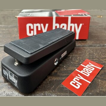 Černý wah-pedál s texturovaným povrchem a gumovou stoupací plochou. Na boku je bílý nápis „cry baby“ a v pozadí je červená krabice s bílým nápisem „c…