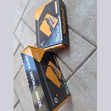 Dvě krabice s produktem BeamZ DMX TRANSCEIVER. Balení jsou černé a oranžové s nápisy „7 UNIVERSES“ a „UNPACK. INSTALL. AMAZE.“. Na krabicích je zobra…