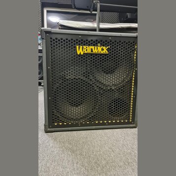 Černý basový reproduktor v šedém rámu s kovovou ochrannou mřížkou. V horní části je žluté logo Warwick. Za mřížkou jsou vidět dva velké reproduktory…
